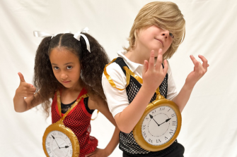 Girl with black hair in bunches and blonde haired boy both wearing 80s tops with watches round their necks